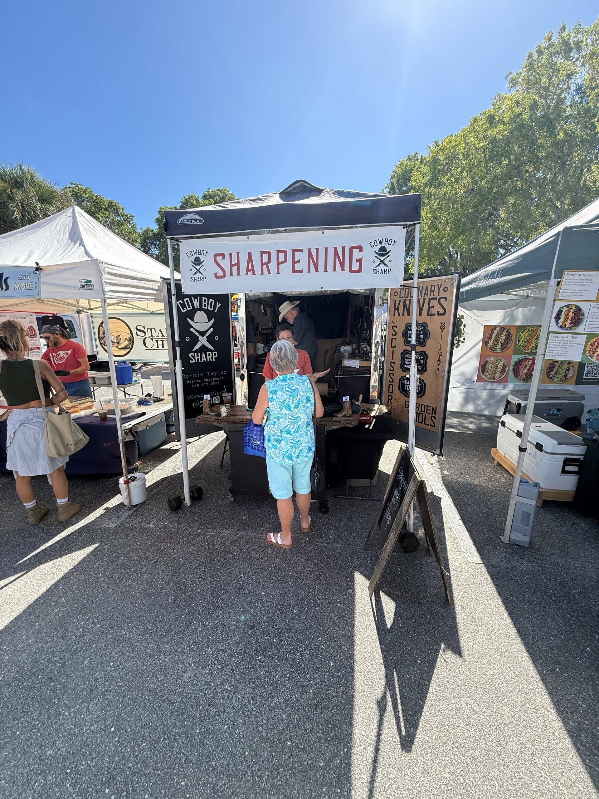 Outdoor market booth labeled “SHARPENING” with a person standing at the counter under bright sunlight.