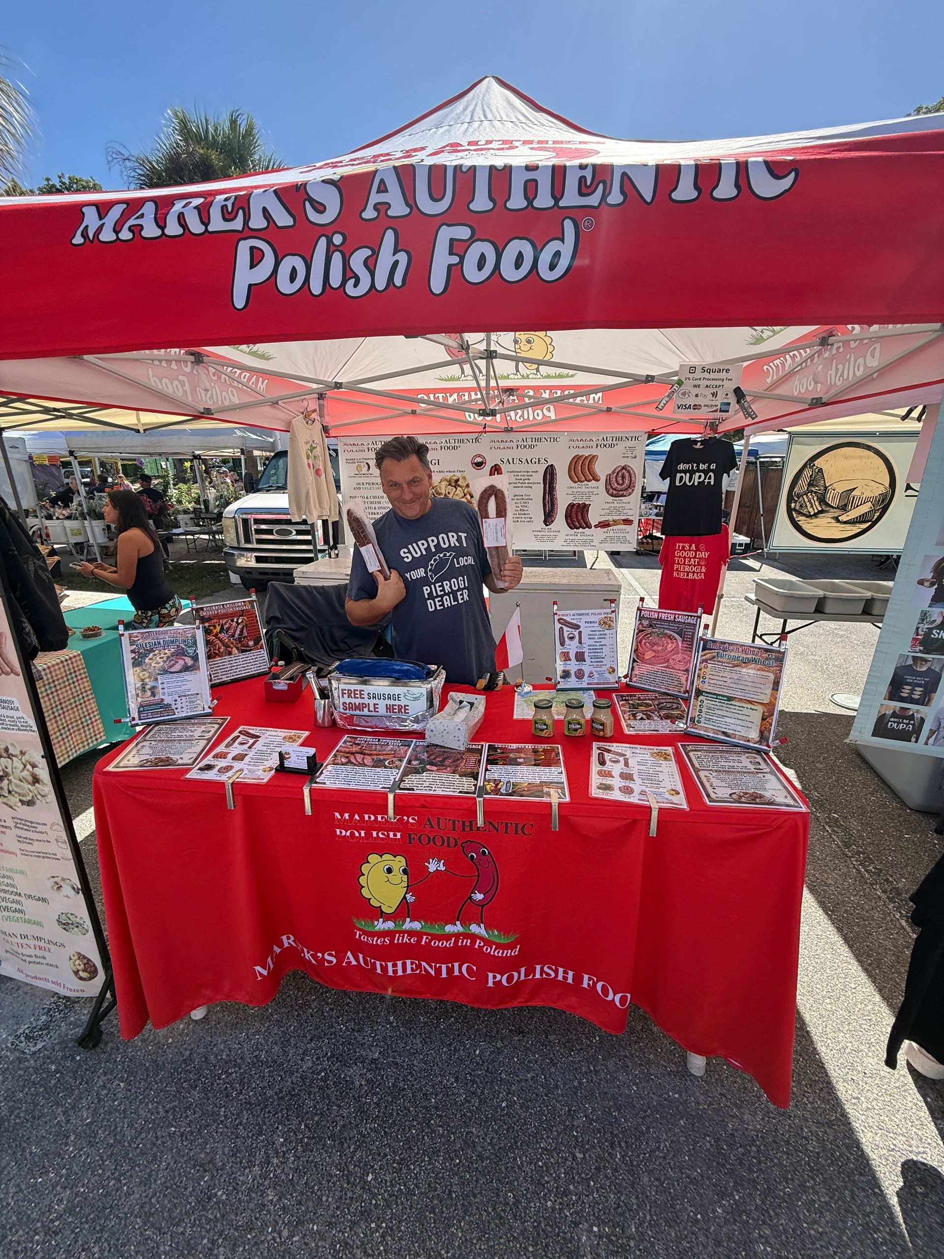 Market stall with red canopy reading “Marek’s Authentic Polish Food,” displaying packaged goods and a vendor behind the table.