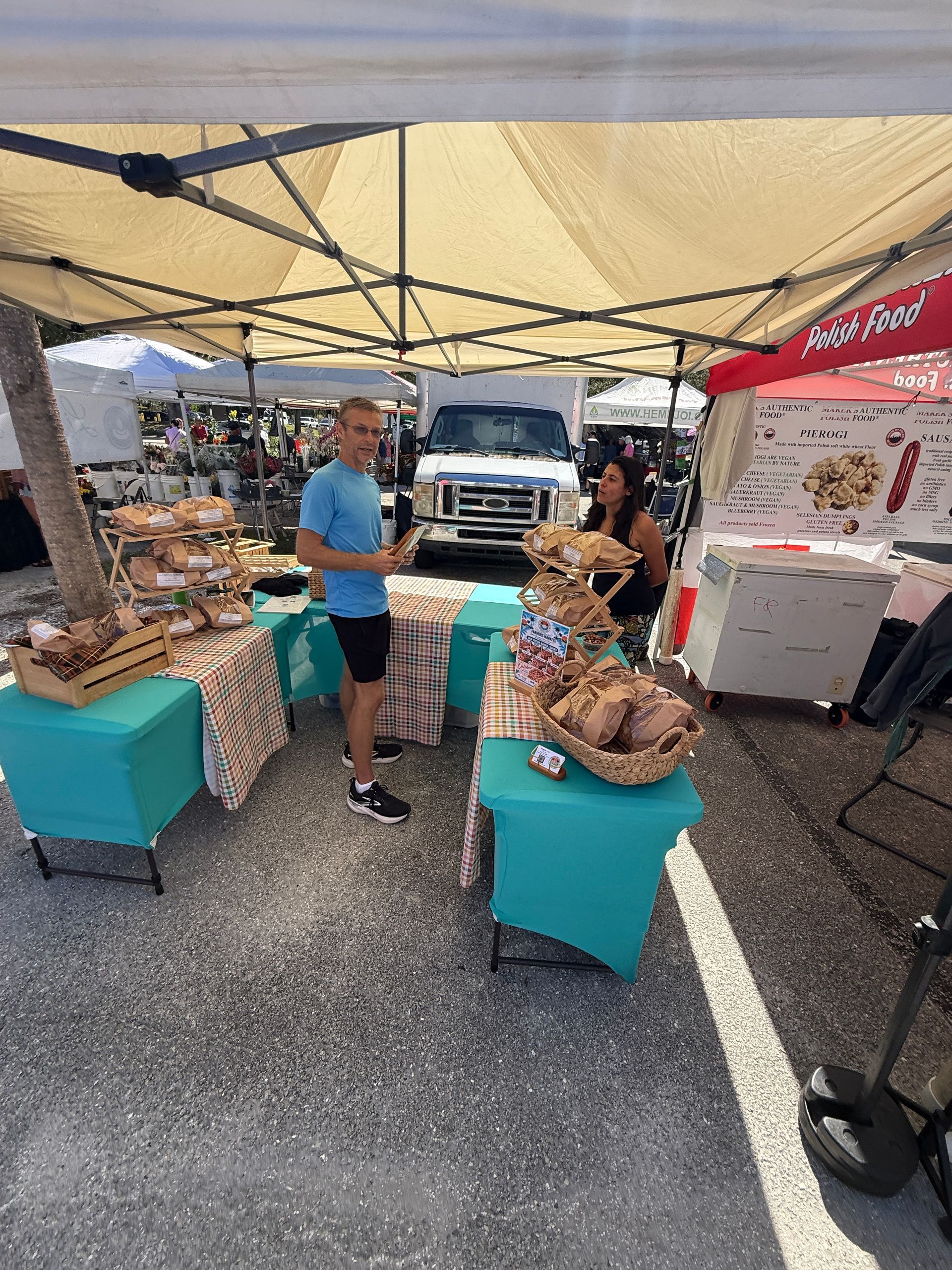 Outdoor market stall under a canopy with baskets of goods and two people talking beside display tables.
