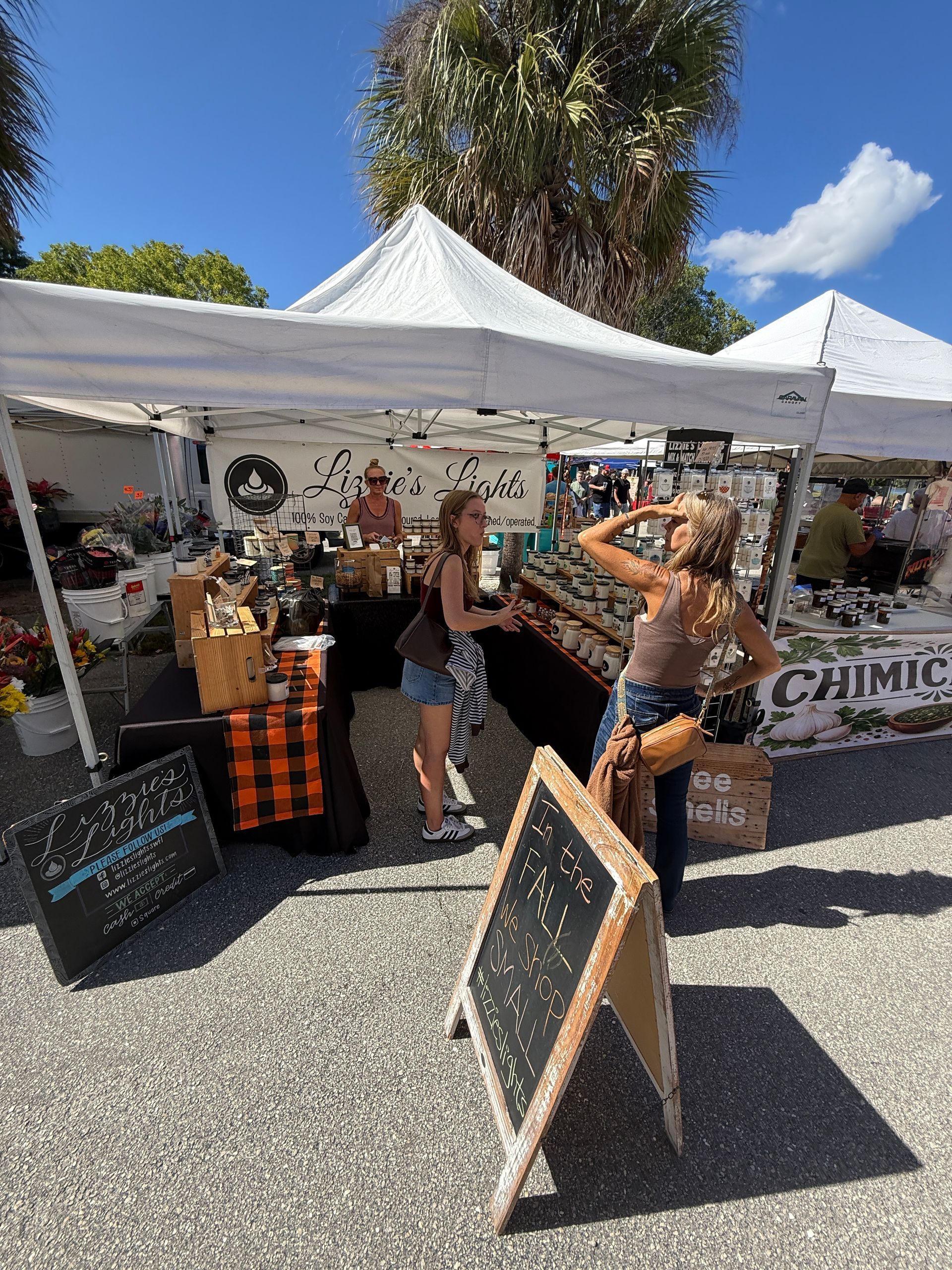 Outdoor market stall with hats and accessories under white tents, with a chalkboard sign in front.