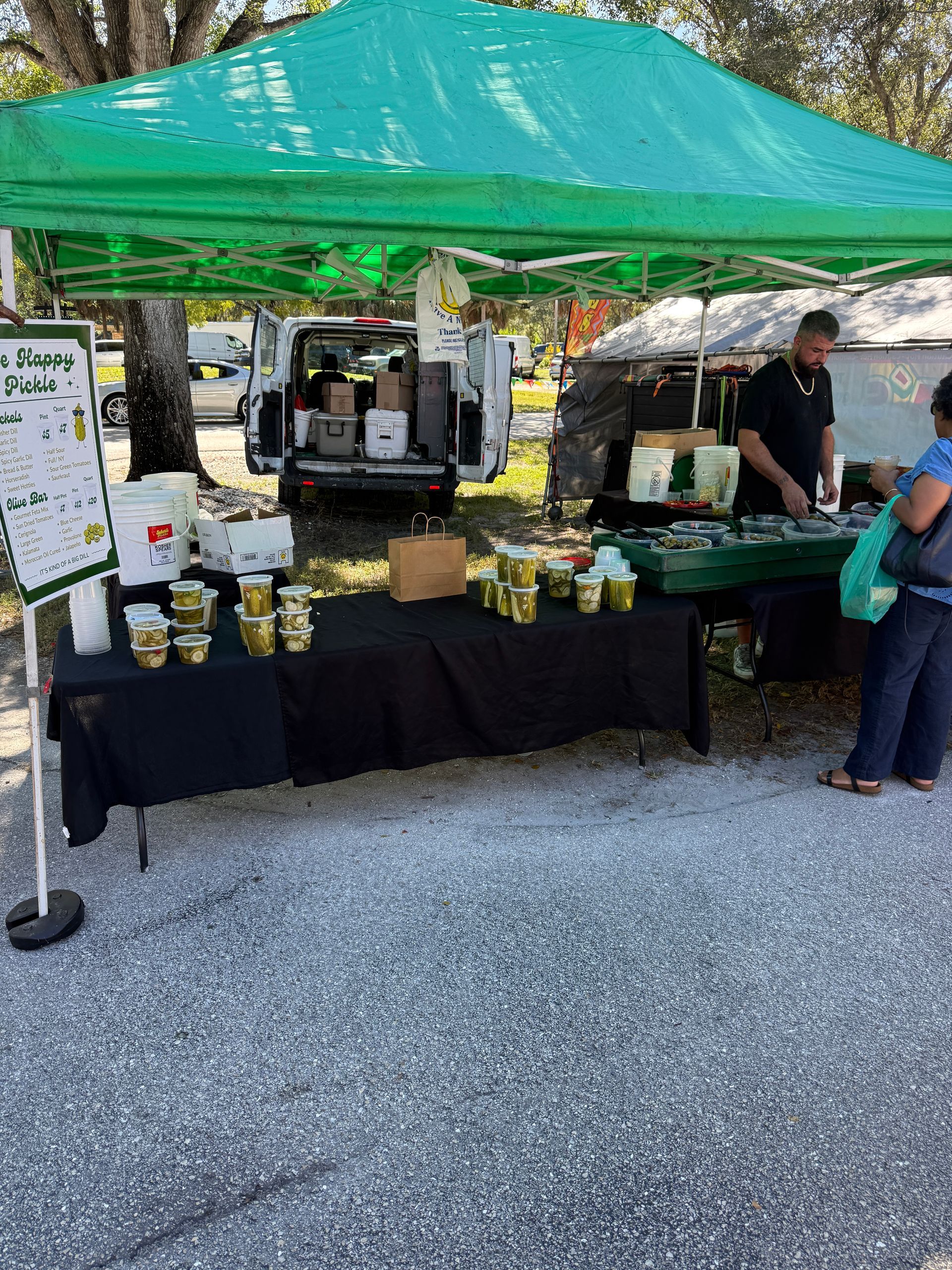Outdoor market stall under a green canopy with bottles, jars, and a vendor serving a customer