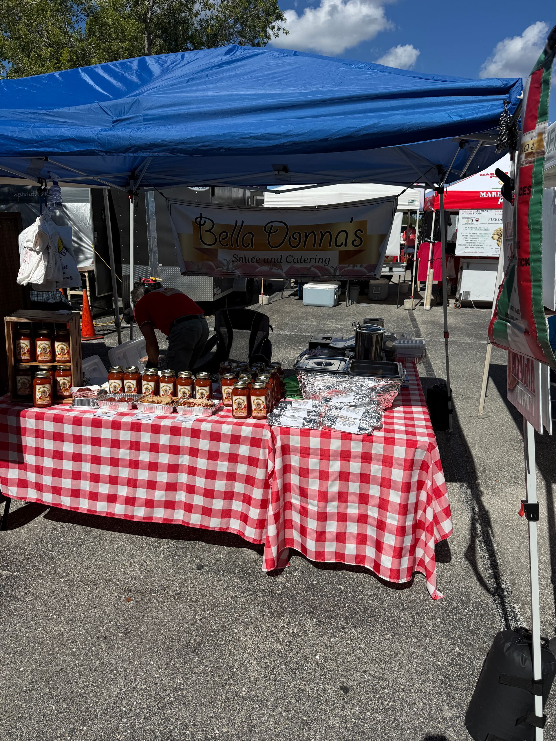 Outdoor market booth with blue canopy, red-and-white checkered tablecloth, and jars and goods on display