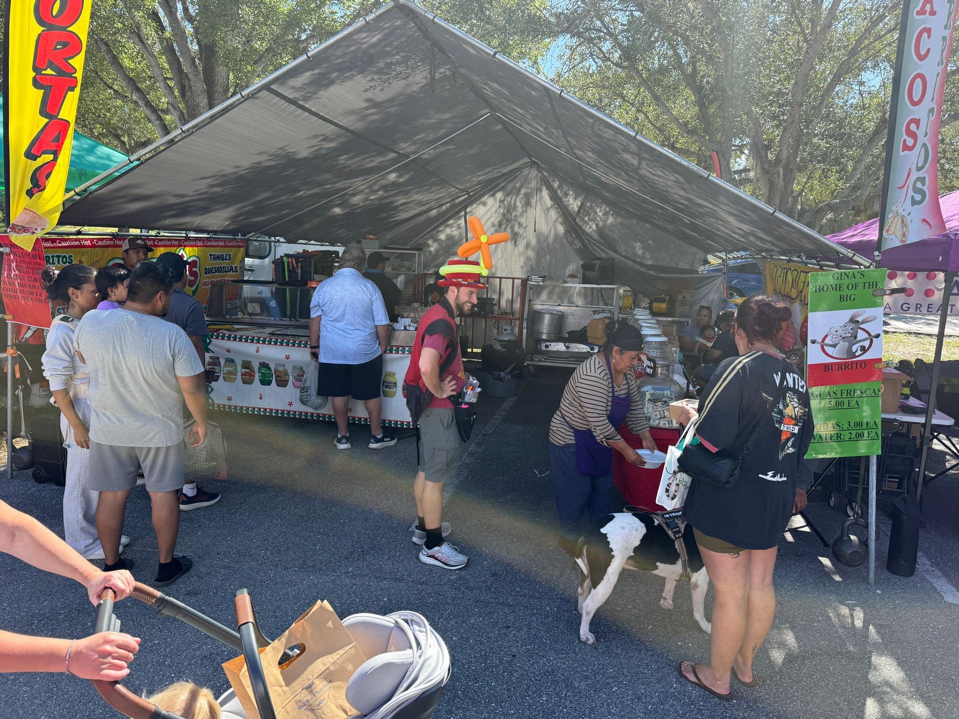 Outdoor market booth under a canopy with people browsing food stalls and a dog on a leash