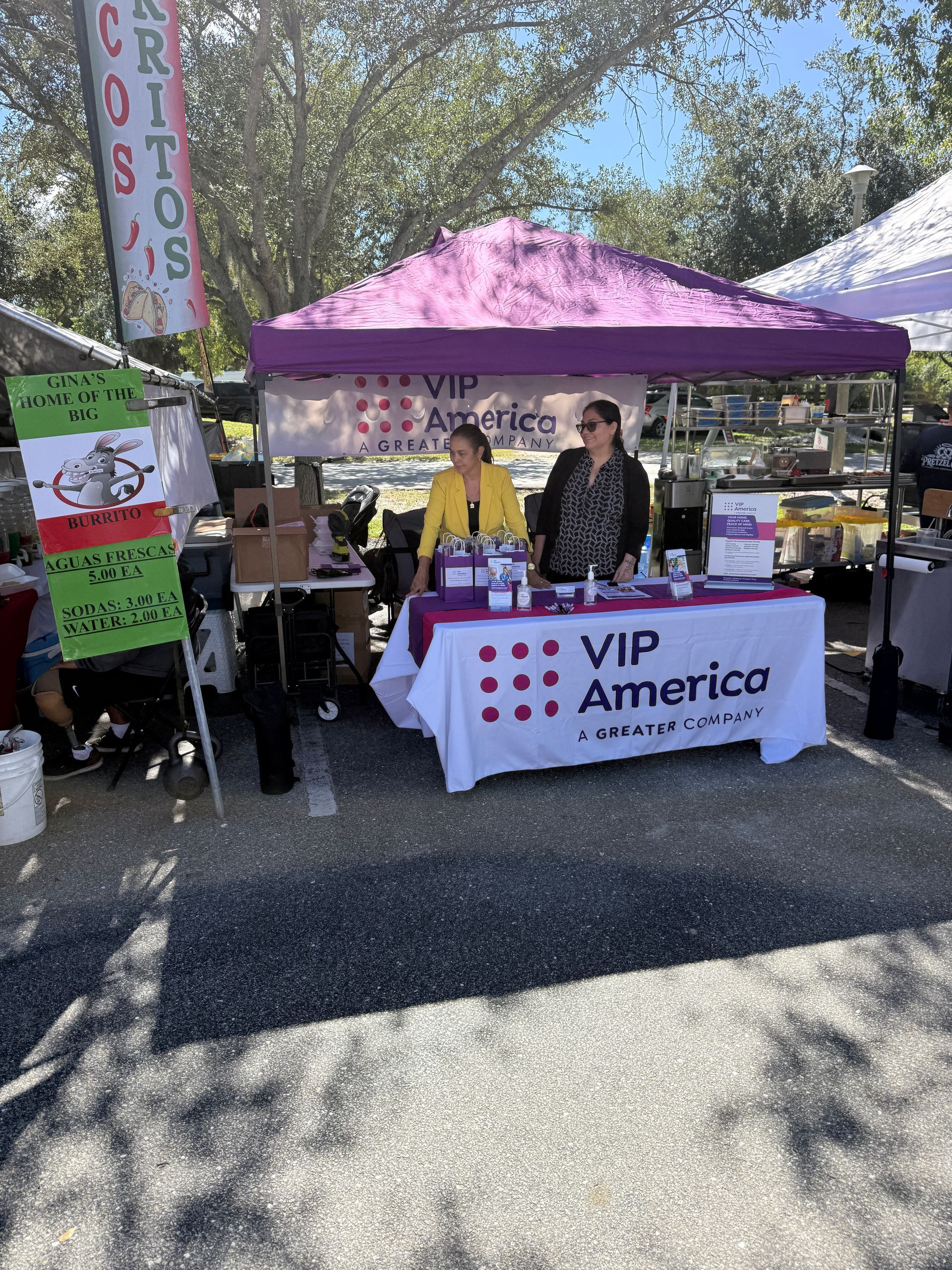 Outdoor VIP America booth with a purple canopy and people behind a table at a sunny market.