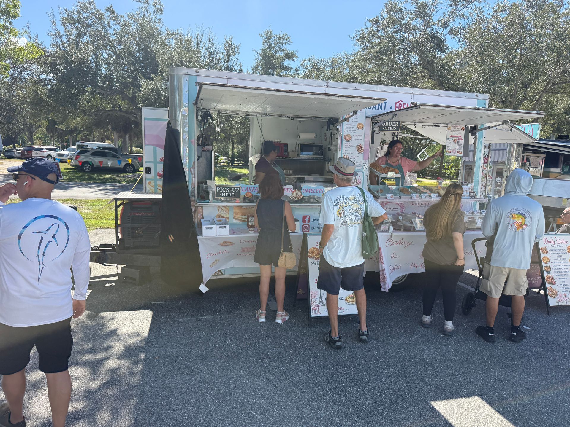 People gathered at an outdoor food truck serving counter in a park on a sunny day