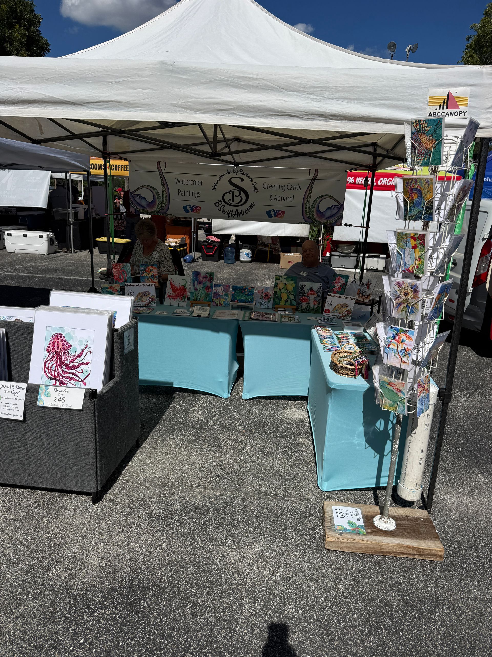 Outdoor craft fair booth under white canopy with art prints and handmade items on display