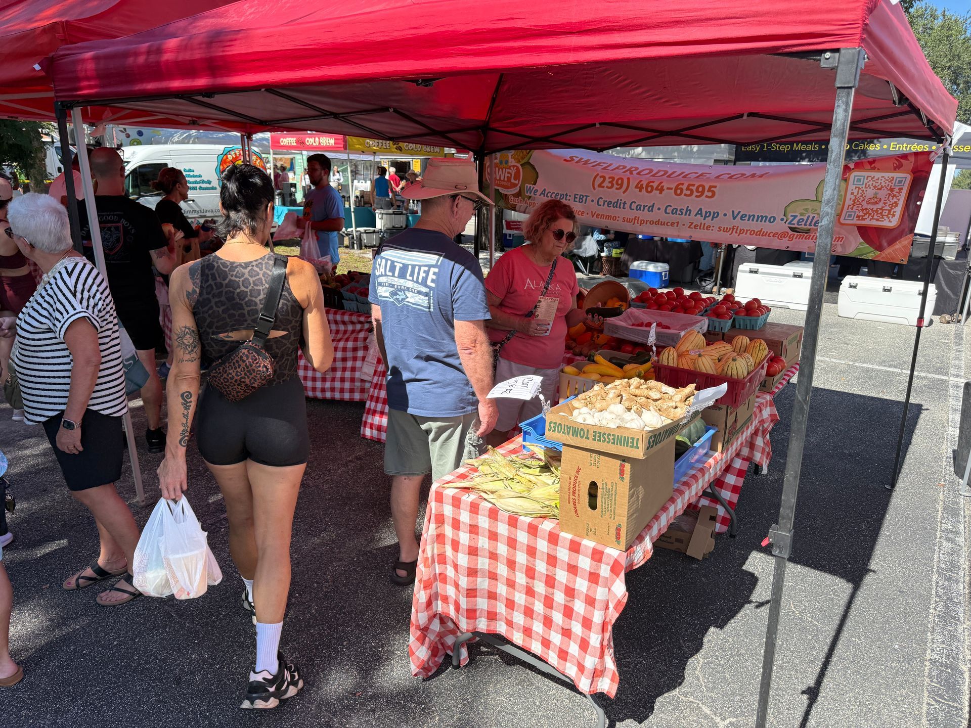 Outdoor market stall under a red canopy with shoppers browsing produce and baked goods