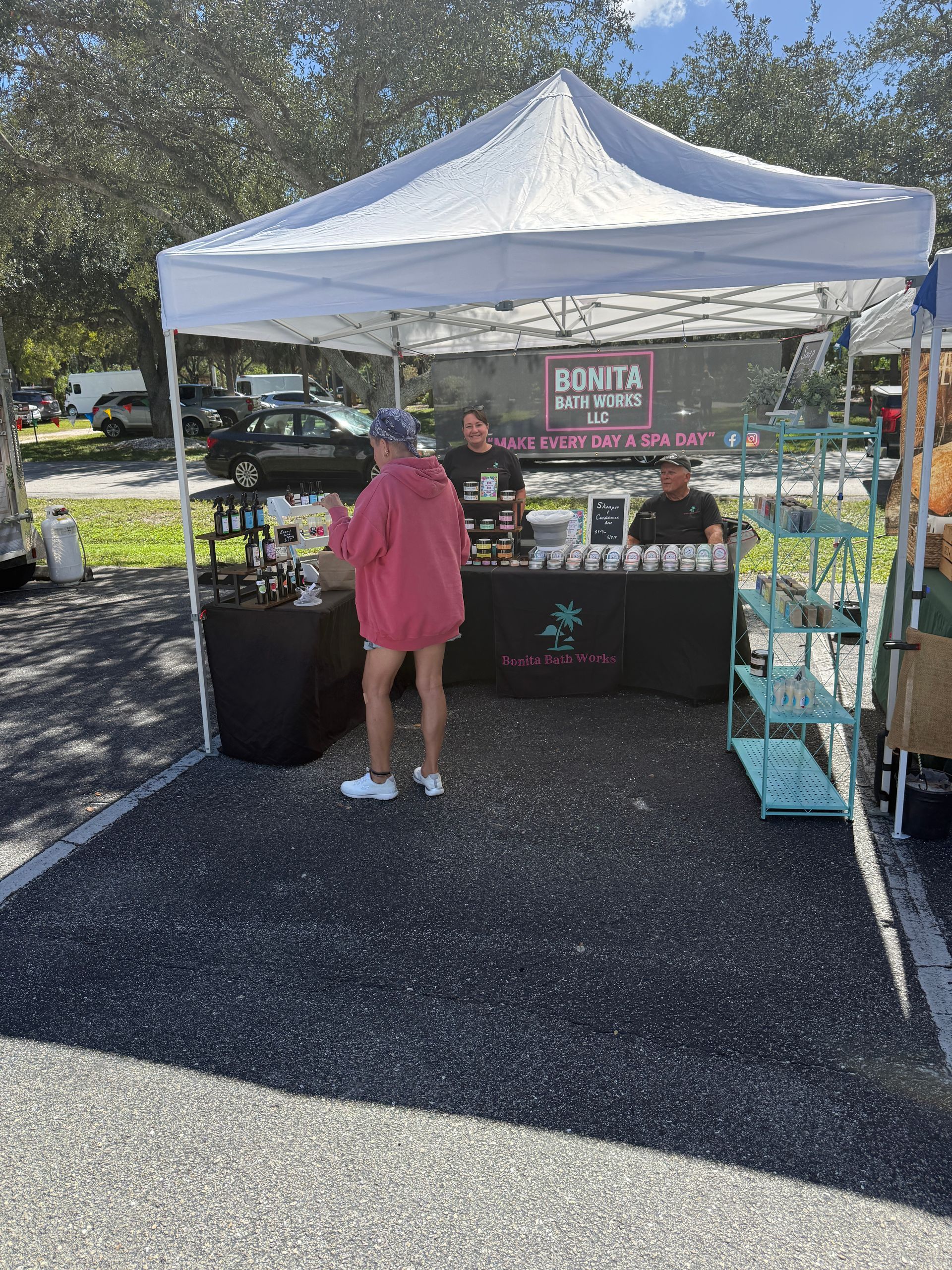 Outdoor market booth under a white canopy, with a person in a pink dress browsing items on display.