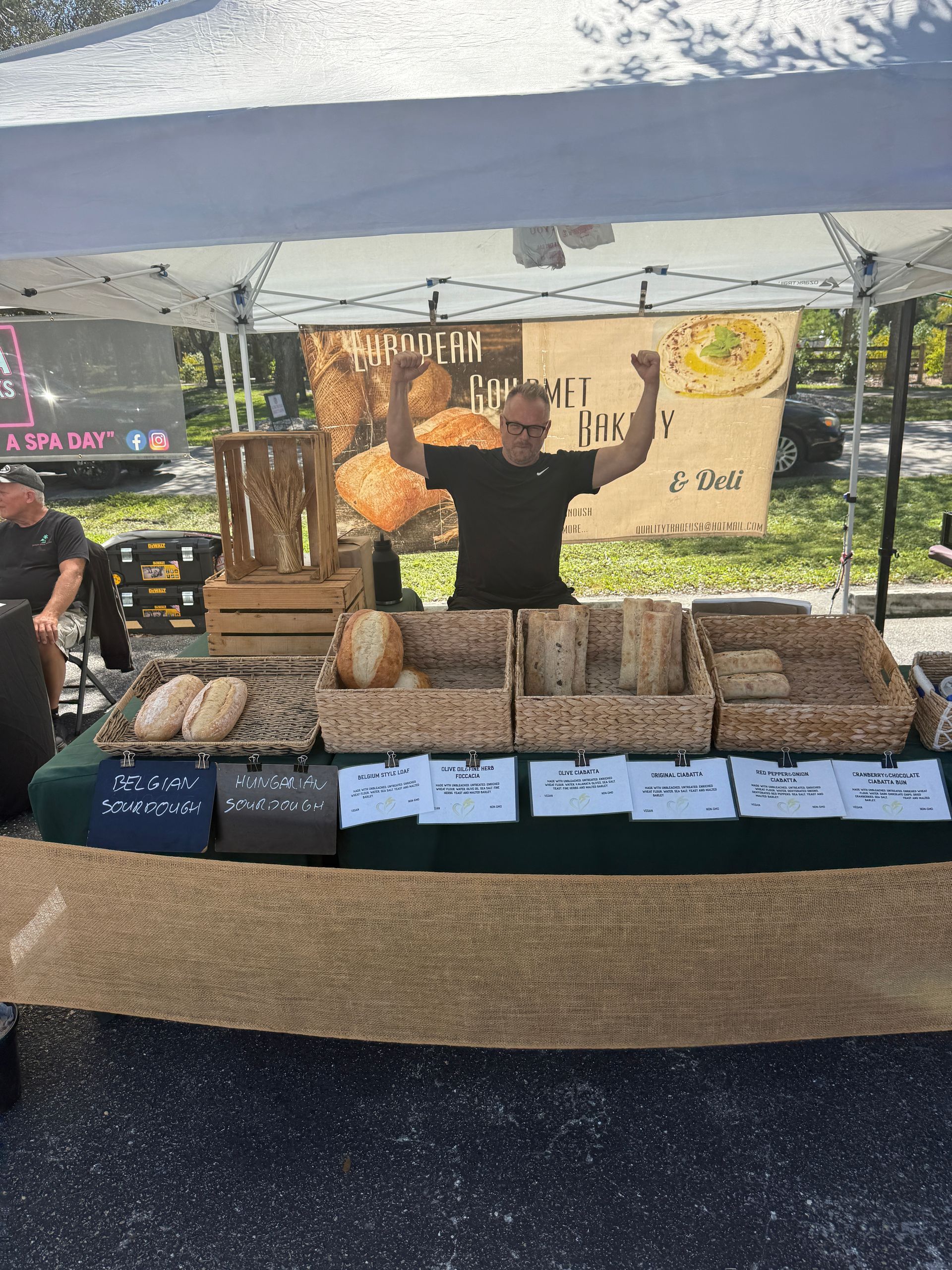 Outdoor market stall with baked goods on display and a vendor standing under a canopy, arms raised.