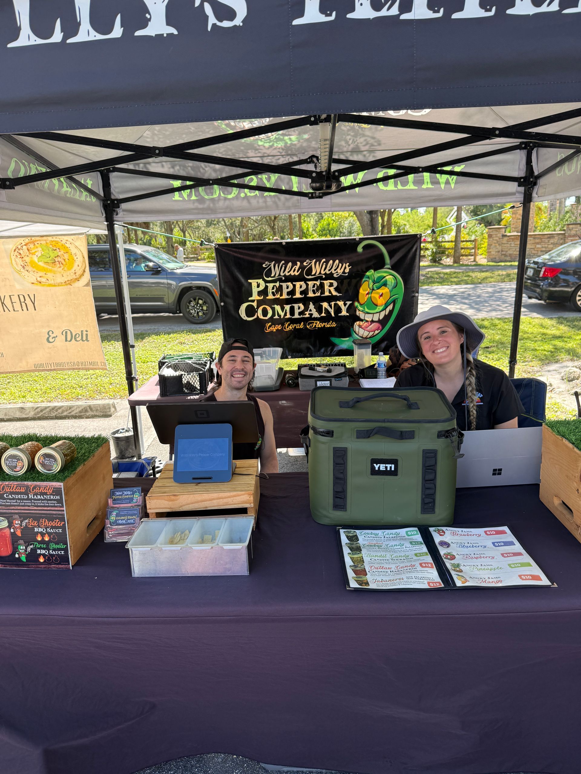 Two vendors at an outdoor market booth with boxes, books, and a cooler under a black canopy.