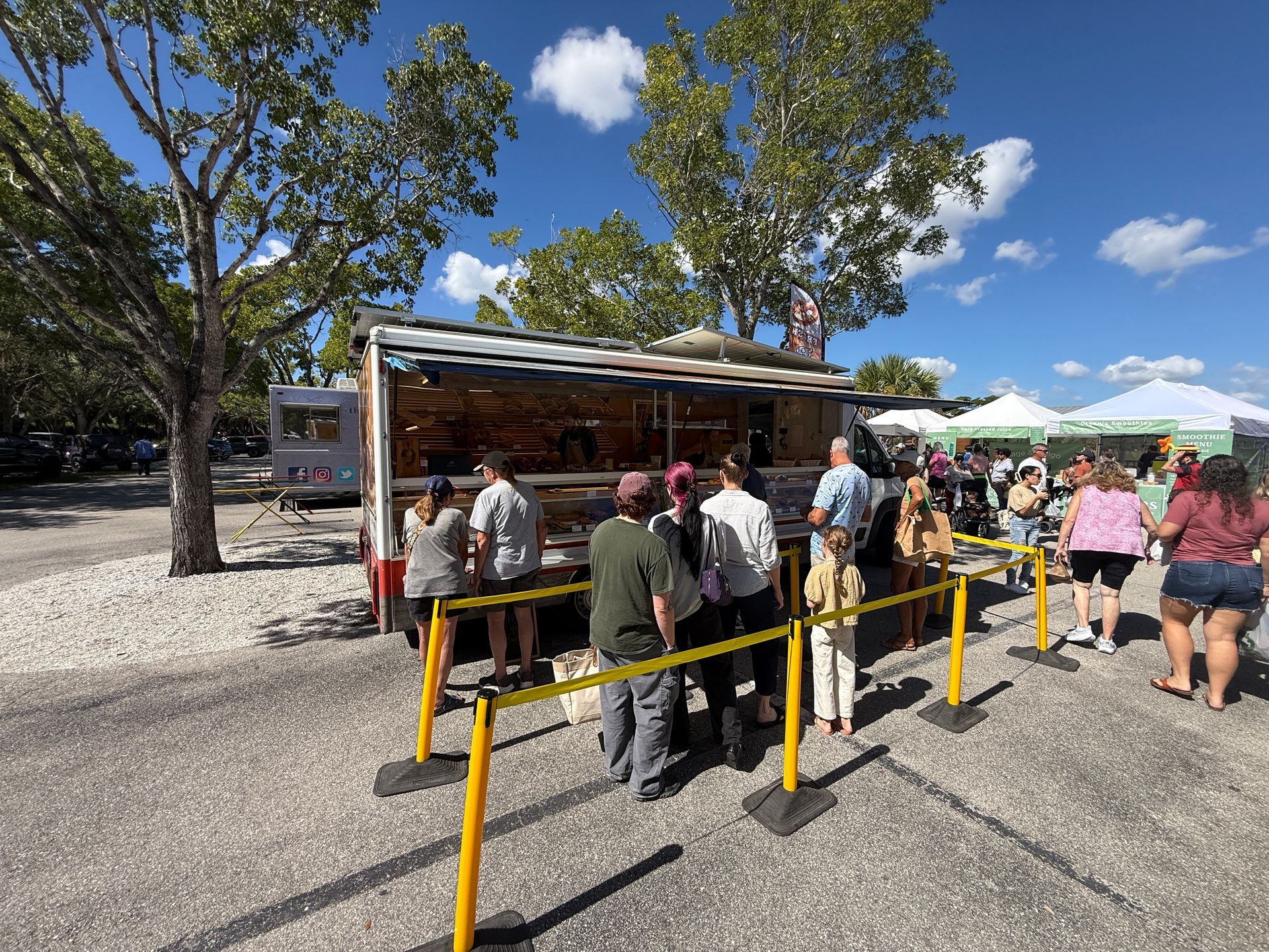 People waiting in line at an outdoor food truck under trees on a sunny day