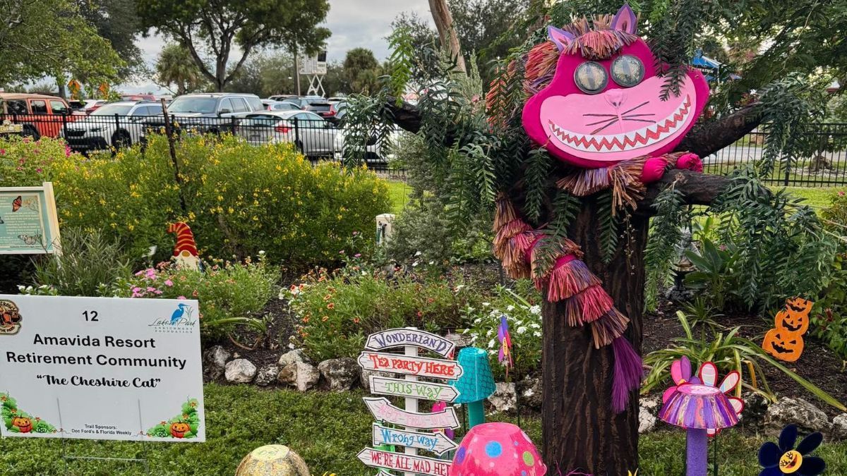 Cheshire Cat display in a garden; pink and purple colors. Cars in the background; seasonal decorations.