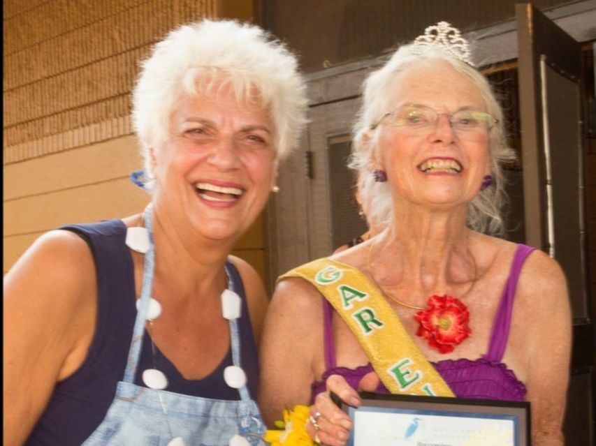 Two older women are posing for a picture and one of them is wearing a sash that says care