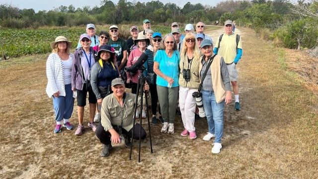 Group of people posing outdoors on a dirt path with binoculars and a telescope, possibly birdwatching.
