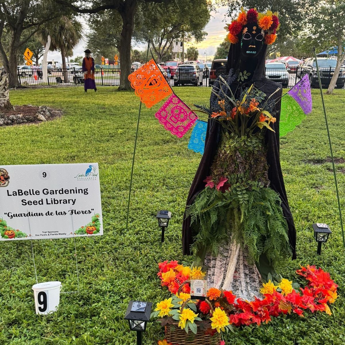 Scarecrow in a garden, decorated with flowers and colorful flags. Text: Labelle Gardening Seed Library.