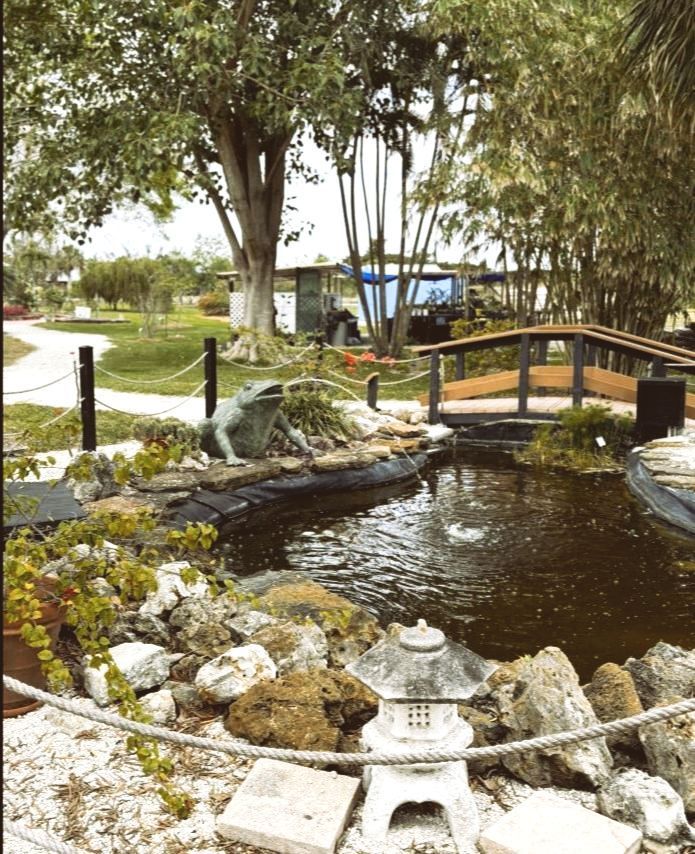 Pond with a small wooden bridge, statue, and Japanese lantern; trees in the background.