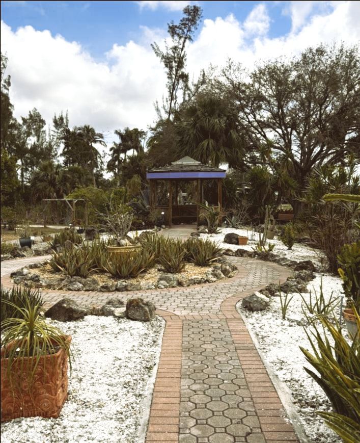 A garden path winds through a botanical garden, leading to a gazebo, under a blue sky.