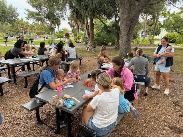People and children sit at picnic tables in a park, possibly engaging in a craft or activity.
