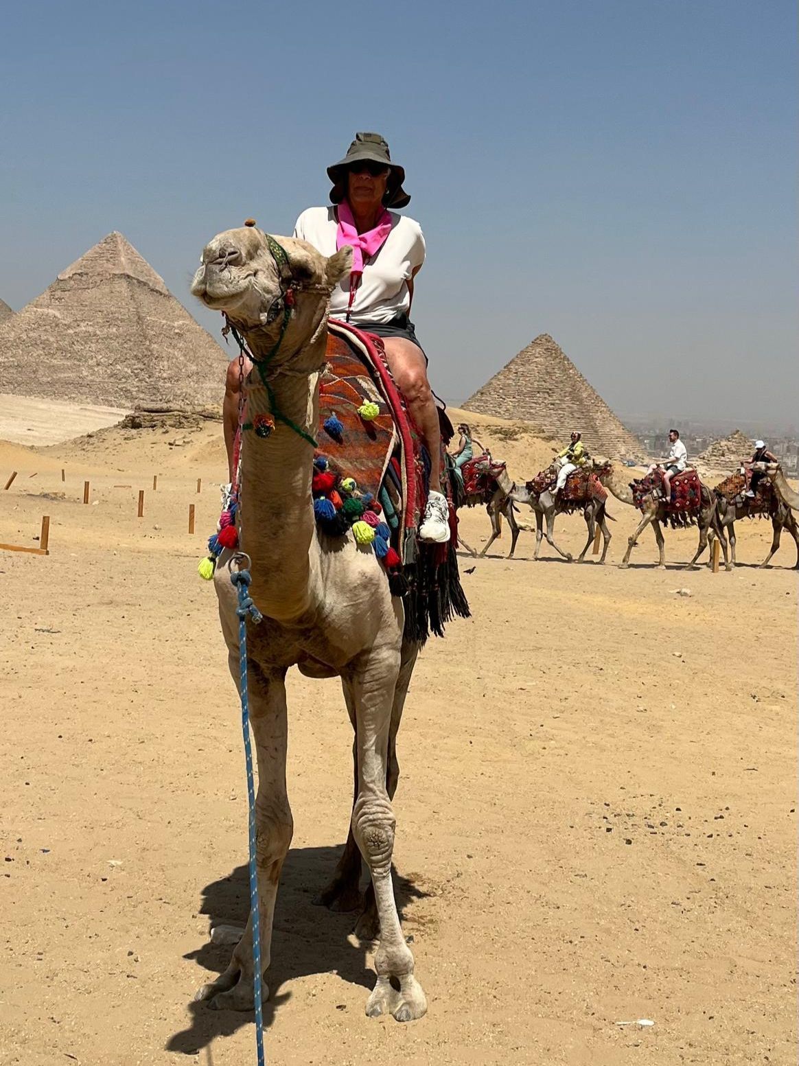 Person rides a camel in front of the pyramids in Egypt, with other camel riders in the background. Sunny day.