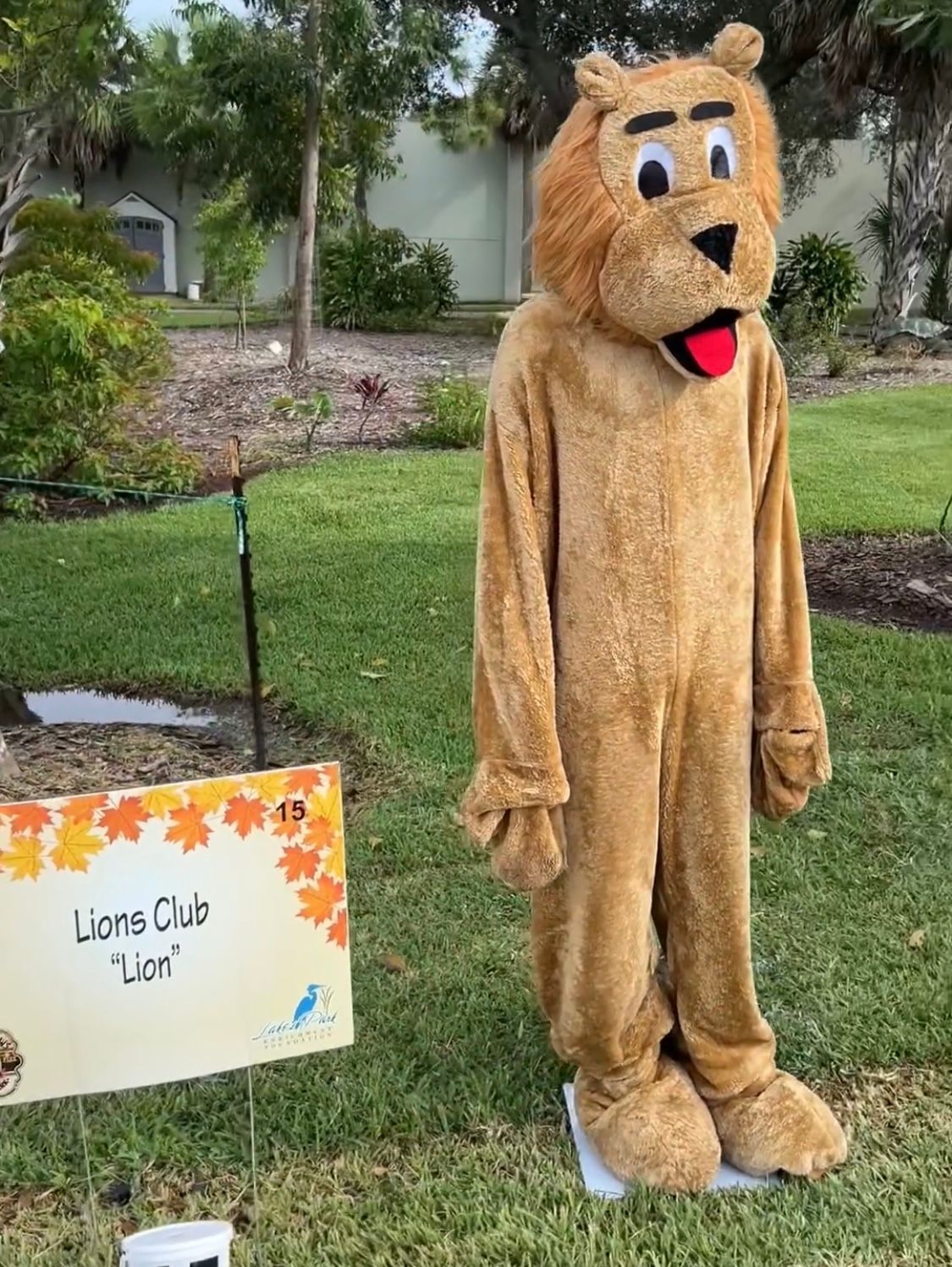 A lion mascot is standing in the grass next to a sign.