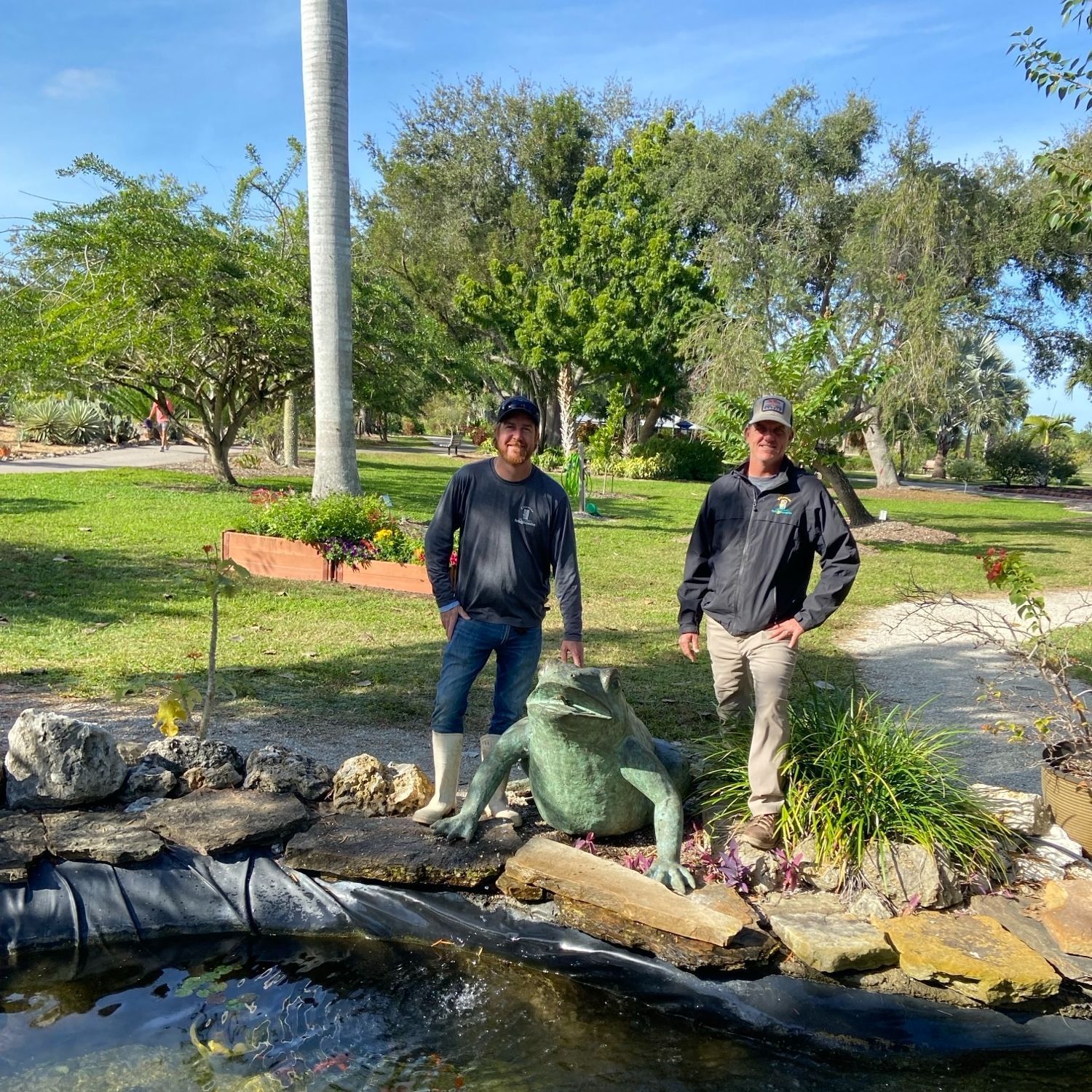 Two men are standing next to a frog statue in front of a pond.