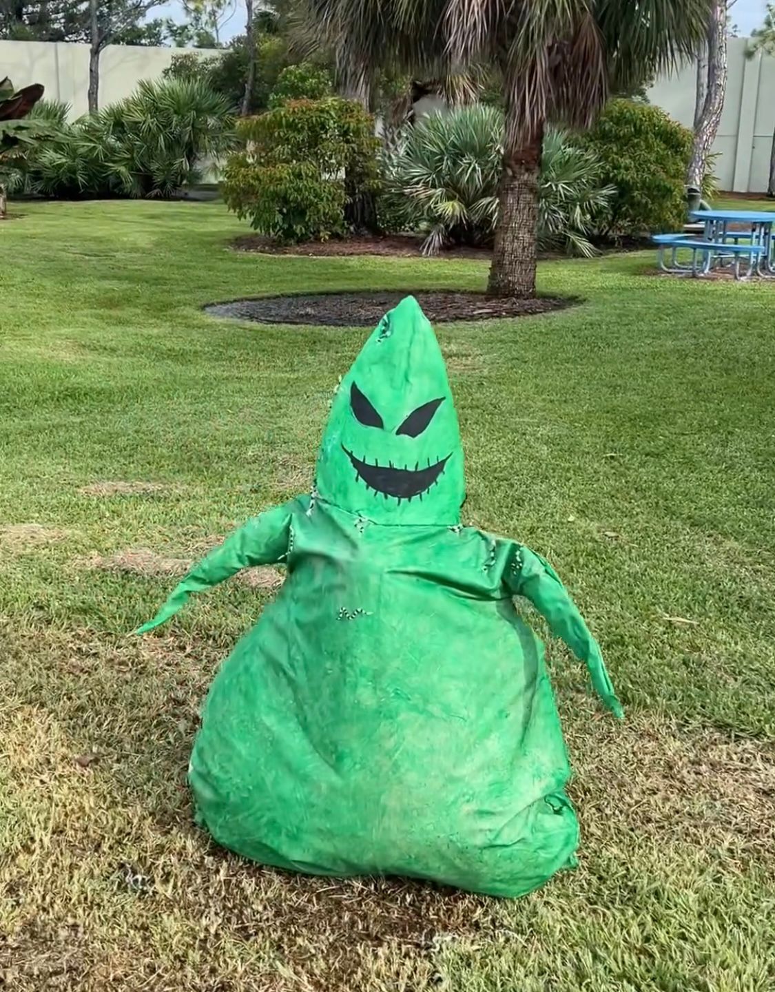A green scarecrow is sitting in the grass in a park.