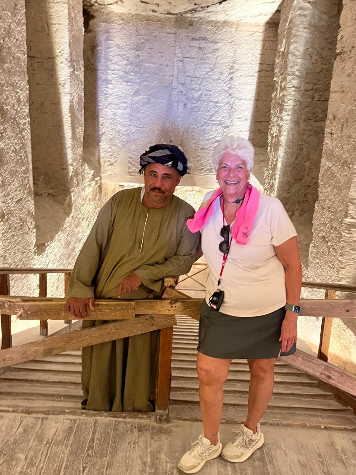 Woman and man pose inside an ancient tomb. The man wears a turban, the woman a pink scarf. Wooden railing.