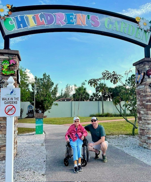 A woman in a wheelchair is posing with a man in front of a children 's garden sign