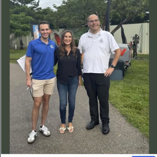 Three people pose outdoors near a military vehicle; man in blue, woman in black, man in white.