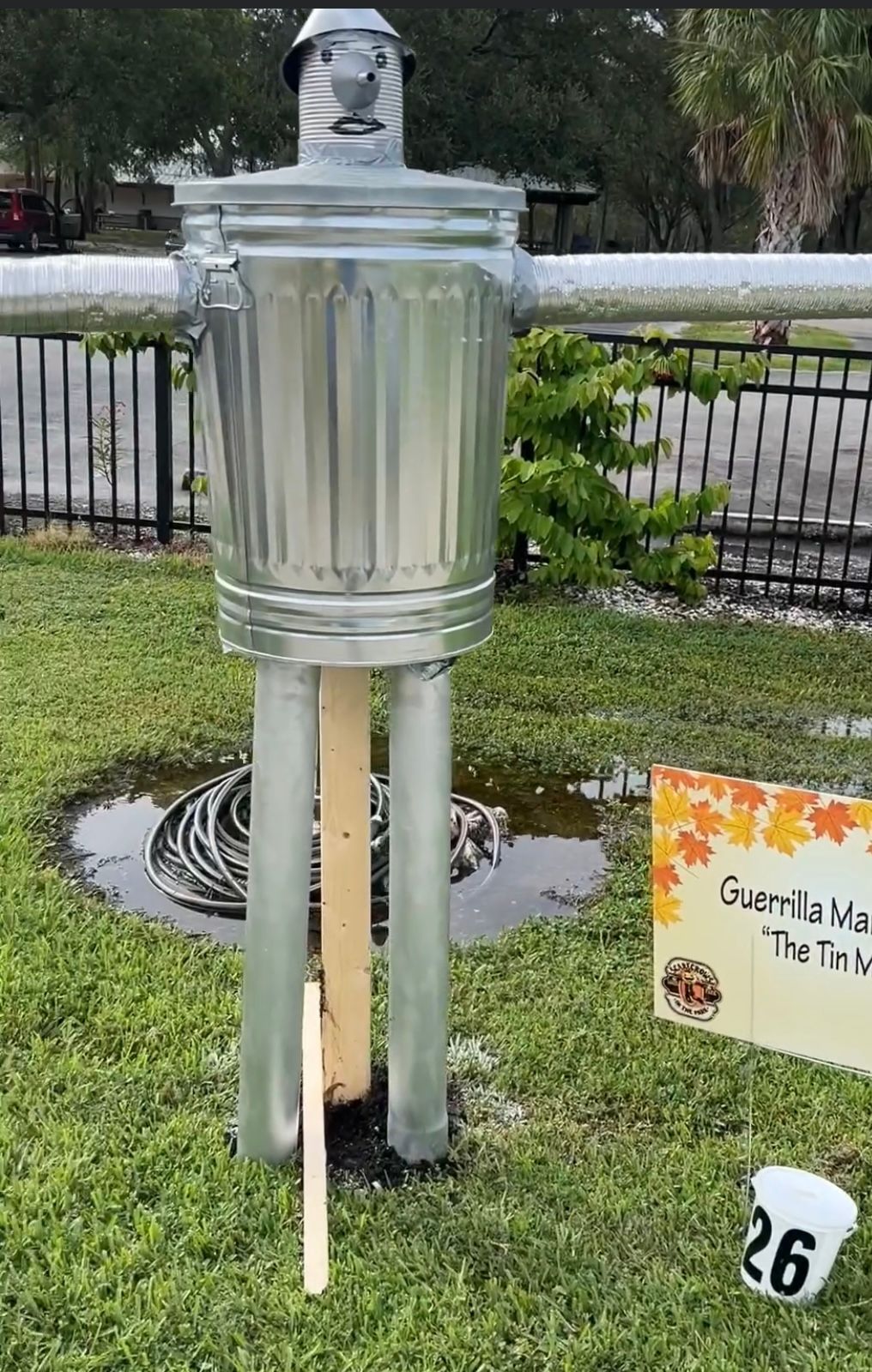 A scarecrow made out of a trash can is standing in the grass next to a sign.