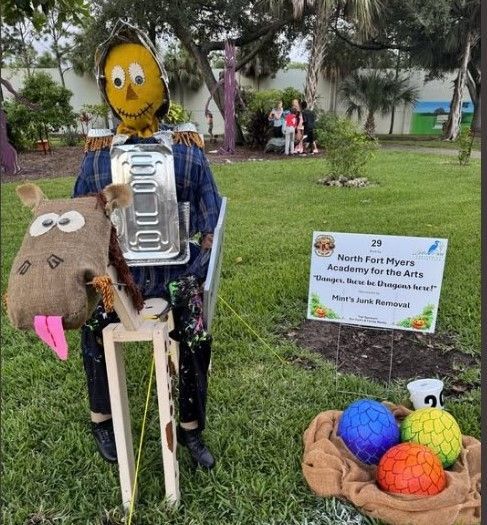 Scarecrow display: a yellow-faced figure with a horse head, holding a sign for an arts academy in a park.