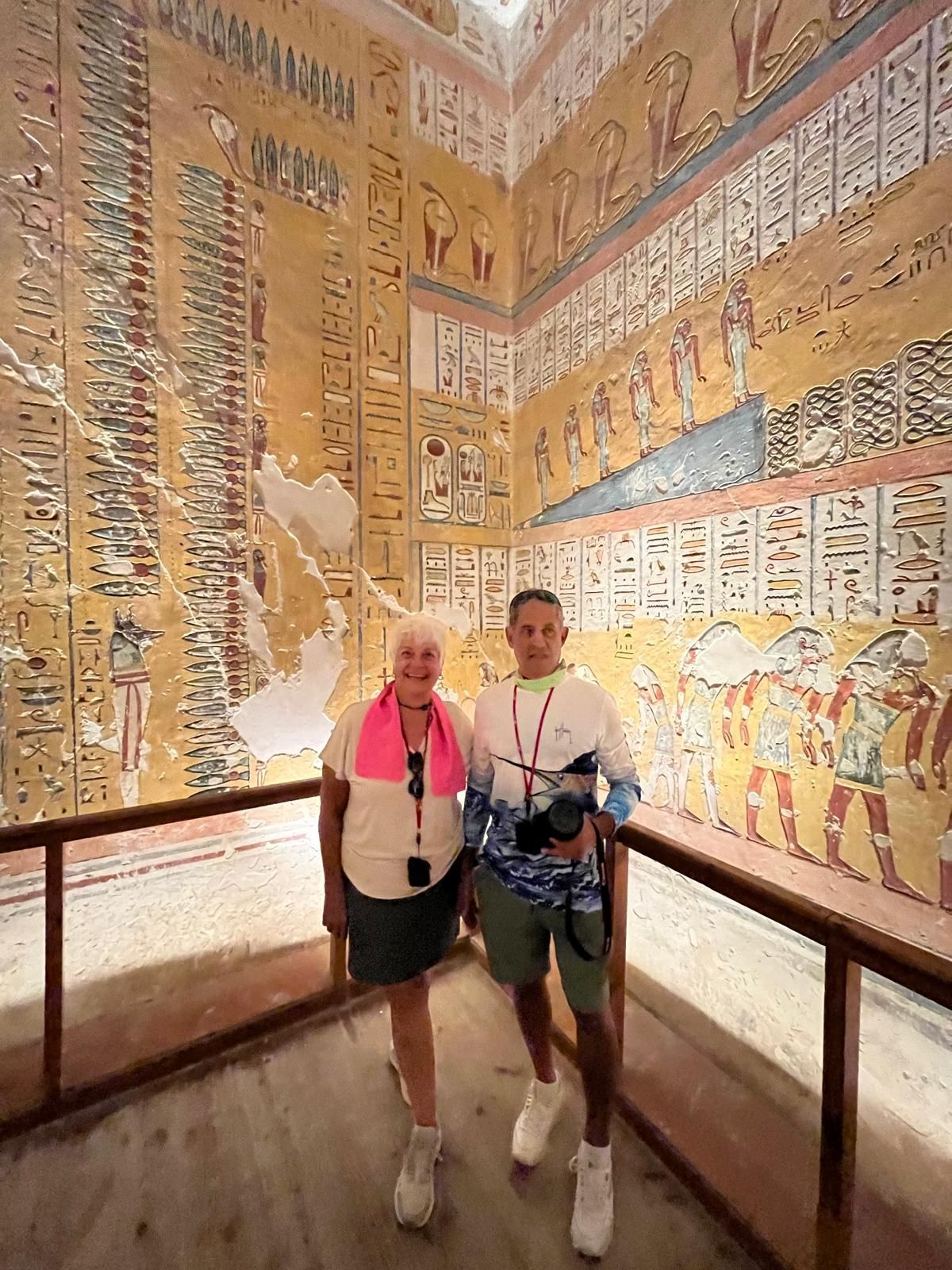 Two people standing in front of ancient Egyptian wall art with hieroglyphs and colorful depictions.