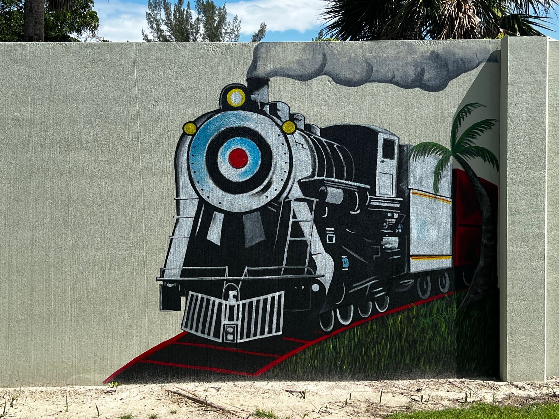 Mural of a black steam train on a gray concrete wall, with red tracks and green grass. Palm tree to the right.