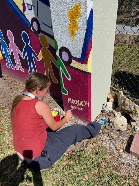 A woman is sitting on the ground painting a mural on a wall with the word mosque on it