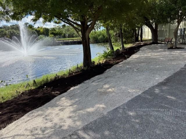 A paved path alongside a lake with a fountain, trees, and newly mulched areas.