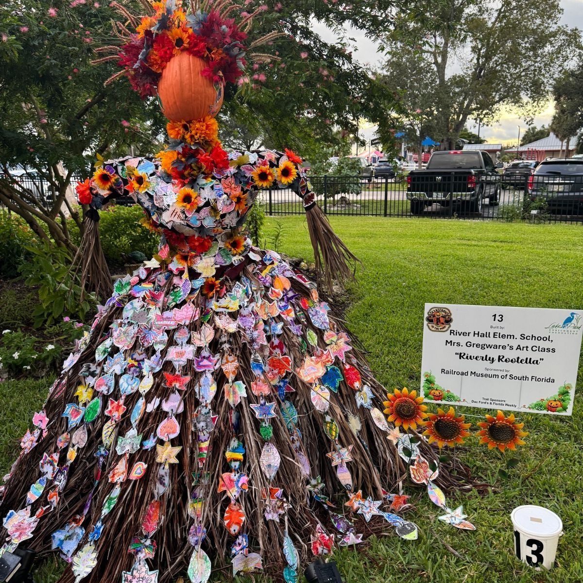Scarecrow with pumpkin head, floral dress, and arms made of branches, on a lawn with a sign.