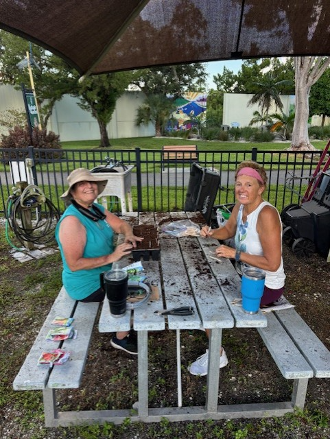 Two people sit at a picnic table, working on something, under a shaded area.