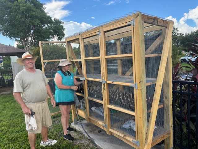 Two people near a large wooden bird coop with wire mesh. The man smiles, the woman opens a door.