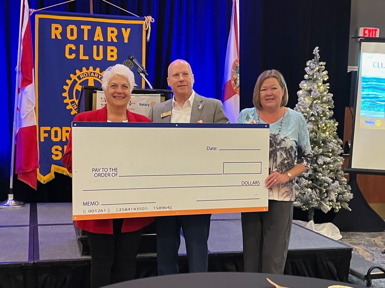 Three people are holding a large check in front of a rotary club banner.