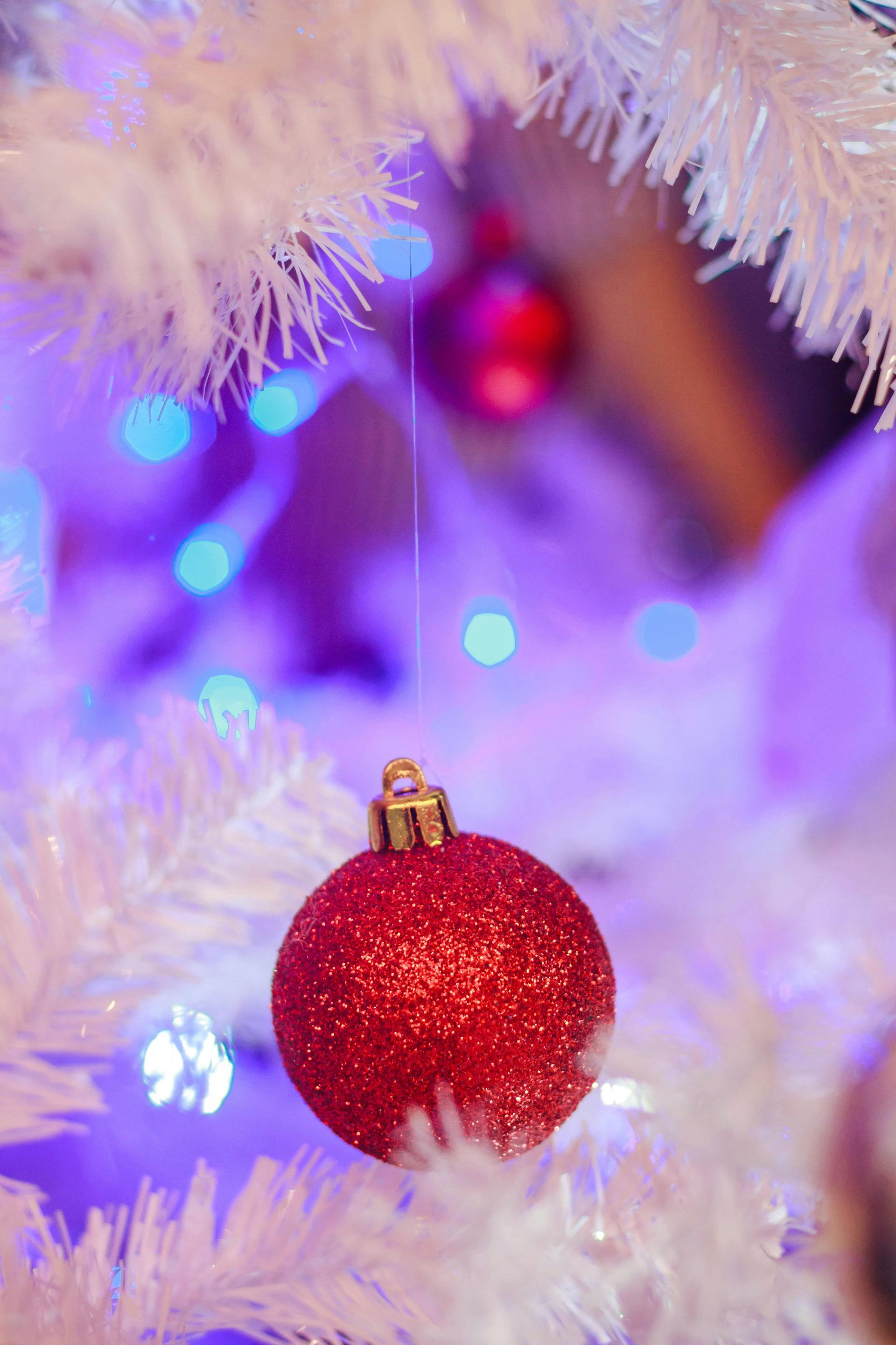 Red sparkly Christmas ornament hanging on a white Christmas tree, with blue and red lights in the background.