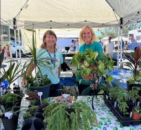 Two women are standing in front of a table full of potted plants.