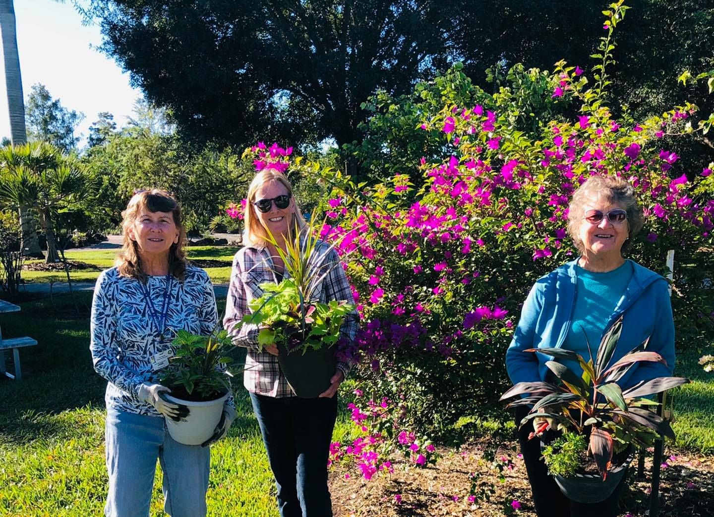 Three women are holding potted plants in front of a bush with purple flowers.
