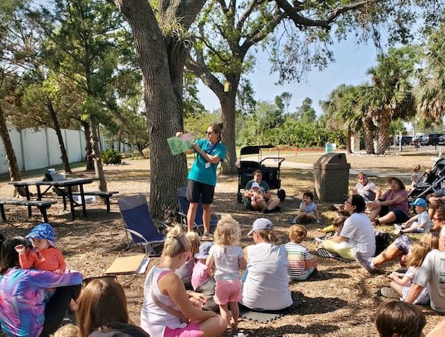 A man is reading a book to a group of people sitting under a tree.
