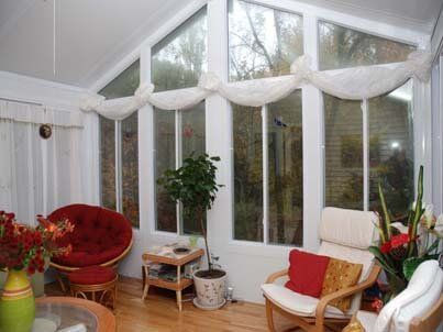 Bright sunroom with white curtains, tall windows, wicker chairs, and a red cushioned chair by plants