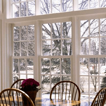 Sunlit dining room with large windows overlooking snowy trees and a table with chairs and flowers.