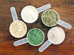 Scoops of protein powders in various colors on a wooden surface.