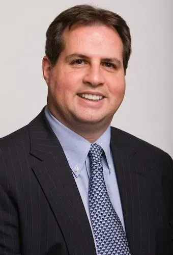 A professional headshot of a smiling man wearing a dark blue pinstriped suit, light blue dress shirt, and patterned tie.