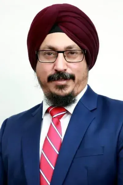 A headshot of a person wearing a maroon turban, glasses, a blue suit, and a red striped tie against a white background.