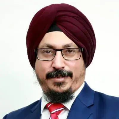 A person wearing a maroon turban, glasses, a blue blazer, and a red-striped tie against a plain white background.