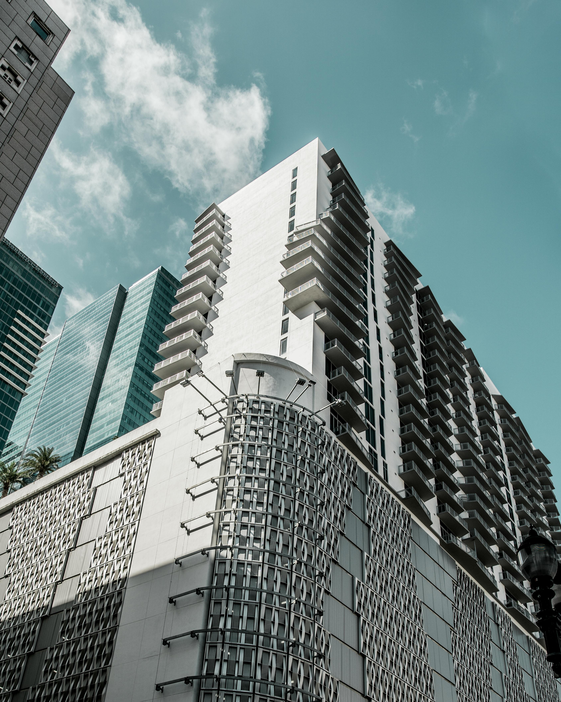 A tall, modern white high-rise building with balcony rows, featuring a decorative textured base against a blue sky.